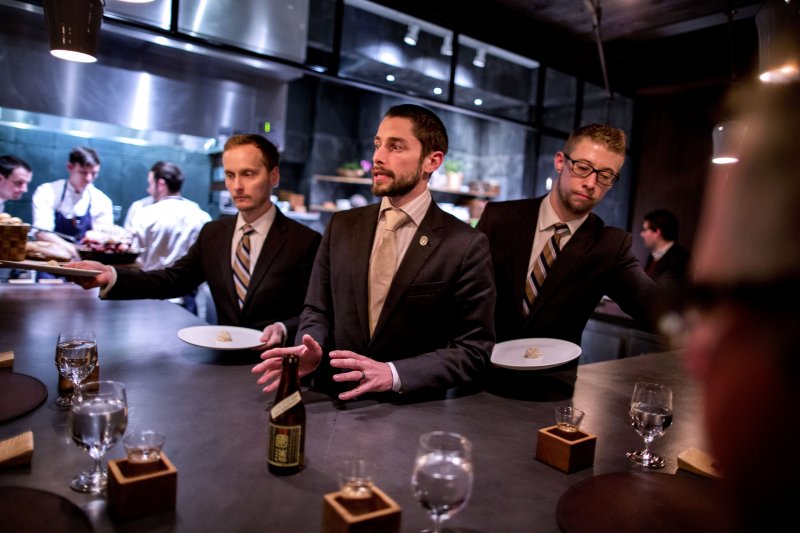 Beverage Director Scott Cameron, center, describes Tentaka Kuni "Hawk In The Heavens" sake during a tasting menu service at Atera in New York, NY on Oct. 26, 2013. Also pictured are waiters Matt Abbick, left, and Joe Giardano, right. The sake was paired with razor clam. A matching drink service pairs various alcoholic beverages with each course, and Cameron describes each beverage for the clientele as they are served.